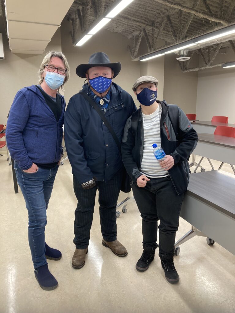 Three people pose for the camera standing next to each other in a college classroom. All are carrying book bags and are wearing masks.