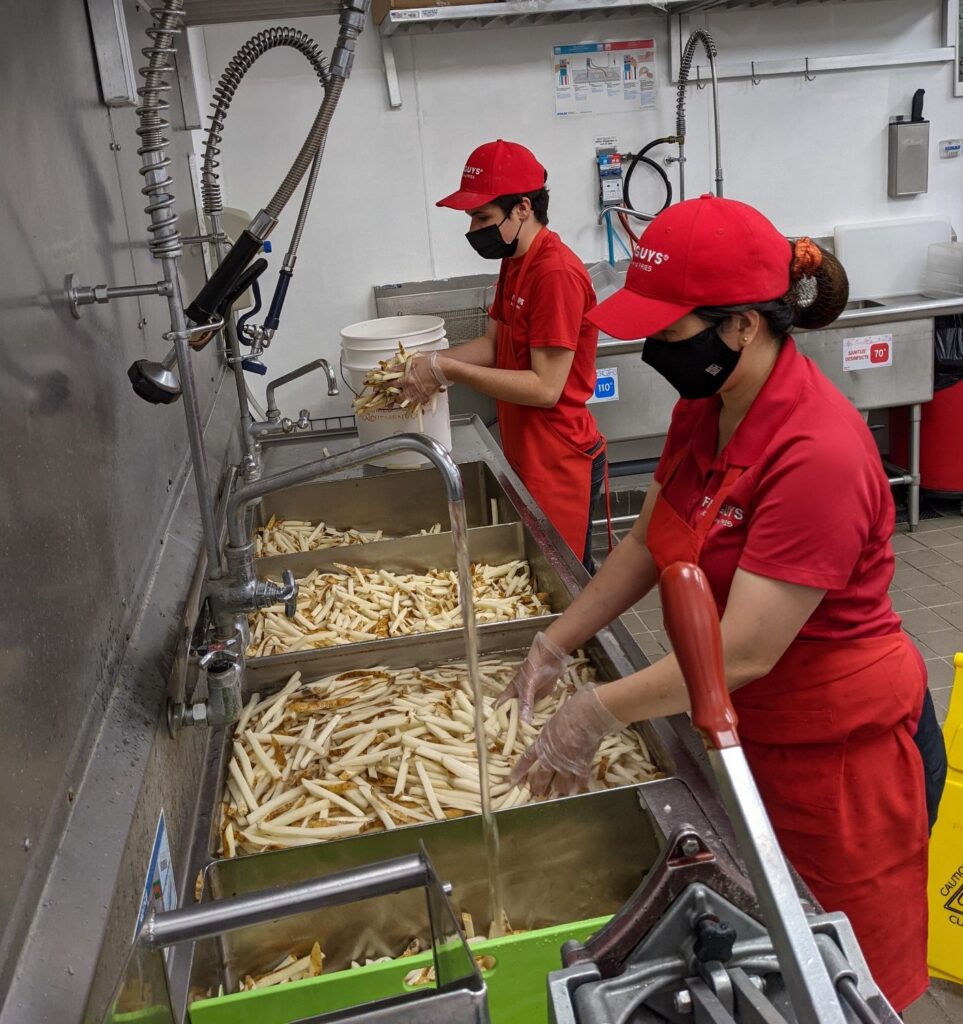 Daniel and a colleague work together at the fry station at 5 Guys. They are wearing red shirts, pants and hats. 