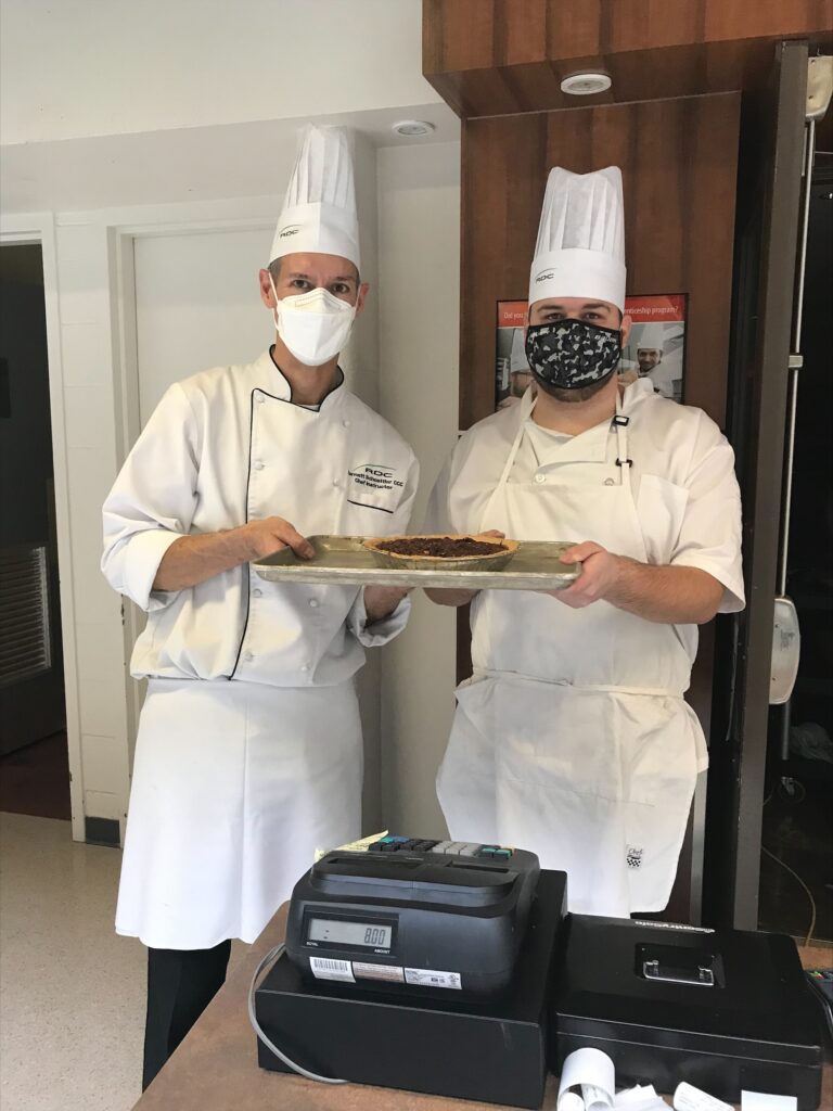 Elijah is on the right, and is standing with his cooking instructor who is to the left of them. They are holding a tray of food together, and are wearing chef's uniforms including white aprons and hats. Both are wearing masks. 