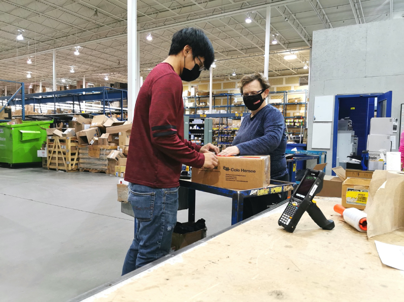 Two employees work at packing a box on a table. They are in a warehouse. The employee on the left is wearing jeans and a red shirt and a black face mask and has short black hair. the employee on the right is wearing a blue shirt and a black mask, and has short brown hair. 