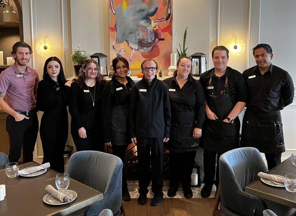 8 employees pose together in a restaurant setting. The 7 on the right are all wearing black uniforms with white name tags pinned to their shirts. The person on the far left is wearing a red shirt. All are smiling.