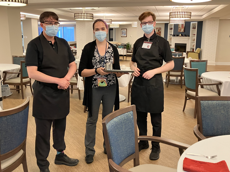 3 service workers pose looking at the camera in a cafeteria setting. They are all wearing black pants, a shirt and blue masks. The middle person is holding a tray of food.