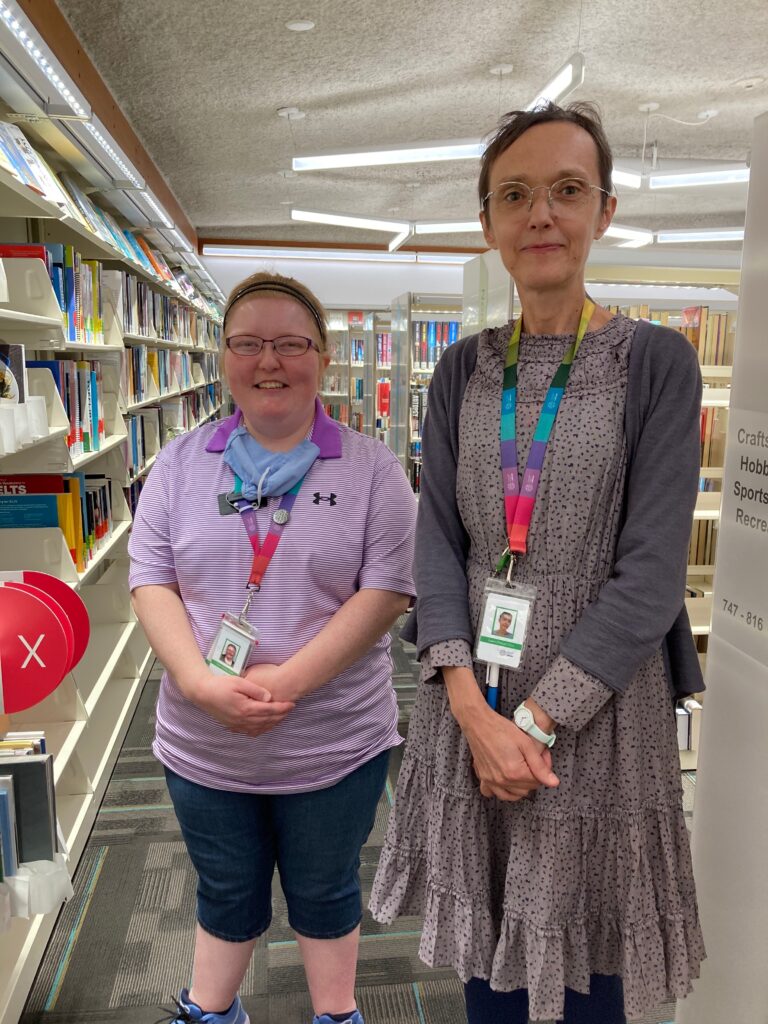 Two library employees pose smiling, with their hands folded in front of them, in front of shelves of books. The person on the left is shorter and is wearing a purple shirt, black shorts and name tag lanyard around their neck. The person on the left is taller, and wearing a grey dress with a grey sweater and a name tag hanging around their neck.