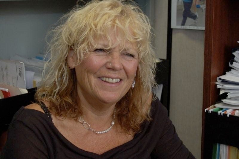 Anne Hughson sits in an office environment, this picture shows her from the shoulders up. She has blonde curly hair and light skin, is wearing a black shirt and a necklace. She is smiling. 